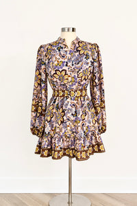 A long-sleeve mini dress with a ruffled hem and waist, featuring an intricate floral and butterfly pattern in purple, gold, and brown, displayed on a mannequin against a white wall.
