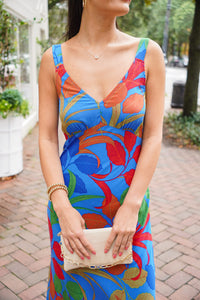 model is wearing a sheath maxi dress in a colorful leaf print with nude heels and is holding a gold purse.