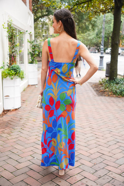 model is wearing a sheath maxi dress in a colorful leaf print with nude heels and is holding a gold purse.