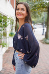 Model is wearing a navy, sheer, cropped top with long sleeves and small embroidered flowers on the top