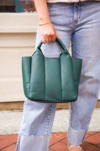 A person wearing light blue jeans holds a dark green pebbled leather handbag with top handles, standing outdoors on a brick surface.
