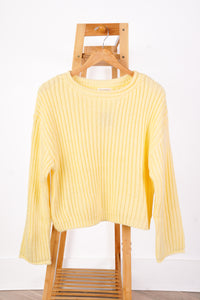 A pale yellow ribbed knit sweater with long sleeves, hanging on a wooden hanger from a wooden step ladder against a white wall.