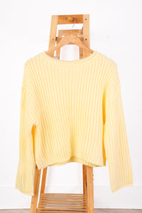 A pale yellow ribbed knit sweater with long sleeves, hanging on a wooden hanger from a wooden clothes rack against a white wall.