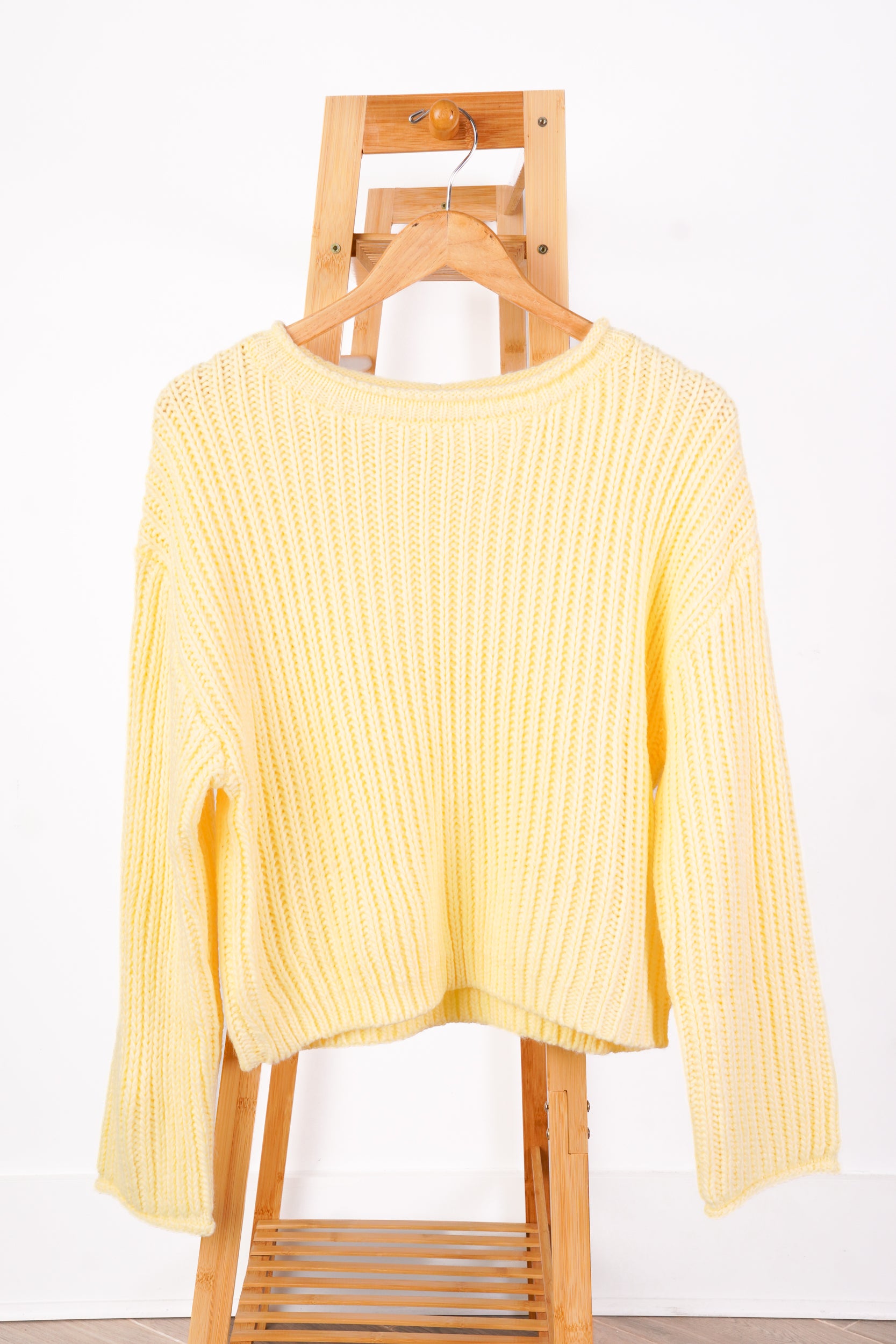 A pale yellow ribbed knit sweater with long sleeves, hanging on a wooden hanger from a wooden clothes rack against a white wall.