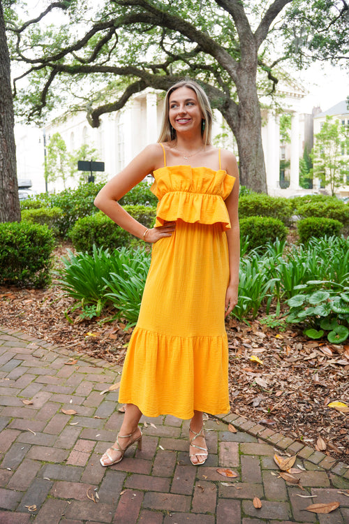Model is wearing an apricot colored midi dress paired with gold heels in front of greenery. 