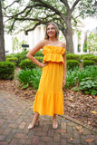 Model is wearing an apricot colored midi dress paired with gold heels in front of greenery. 