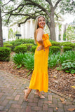 Model is wearing an apricot colored midi dress paired with gold heels in front of greenery.