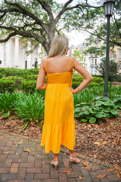 Model is wearing an apricot colored midi dress paired with gold heels in front of greenery.