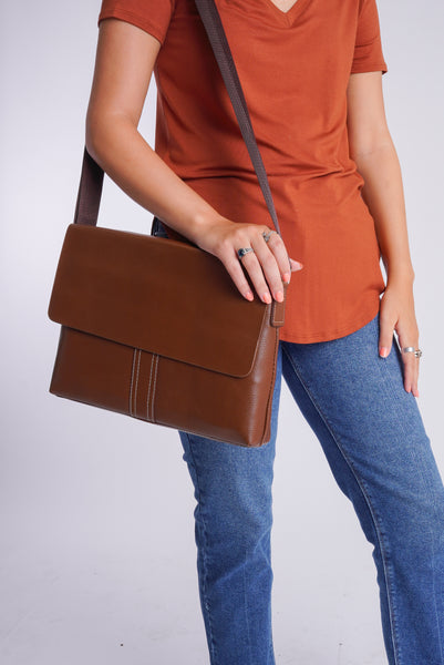 Model is wearing a rust colored tee shirt with a pair of straight leg denim. She is carrying a brown over the shoulder bag with a flap closure and white contrast stitching on brown faux leather. 