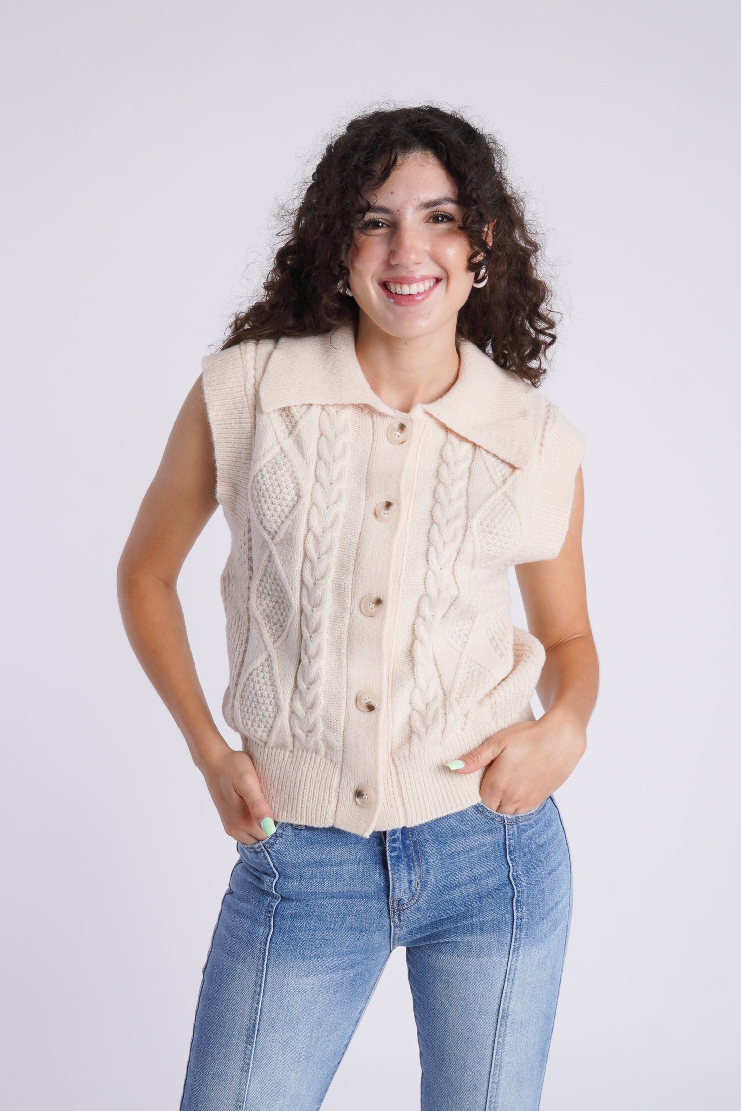 Model is wearing a Cream colored, cable knit sweater vest with a pair of straight leg, seam front denim. 