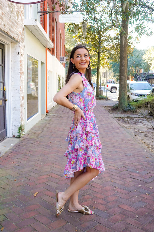 Model is wearing a dress in a floral print in blue, grey, yellow, and pink colors. It is a midi length dress has an asymmetrical skirt hem with tiered and ruffle details, and a high neckline