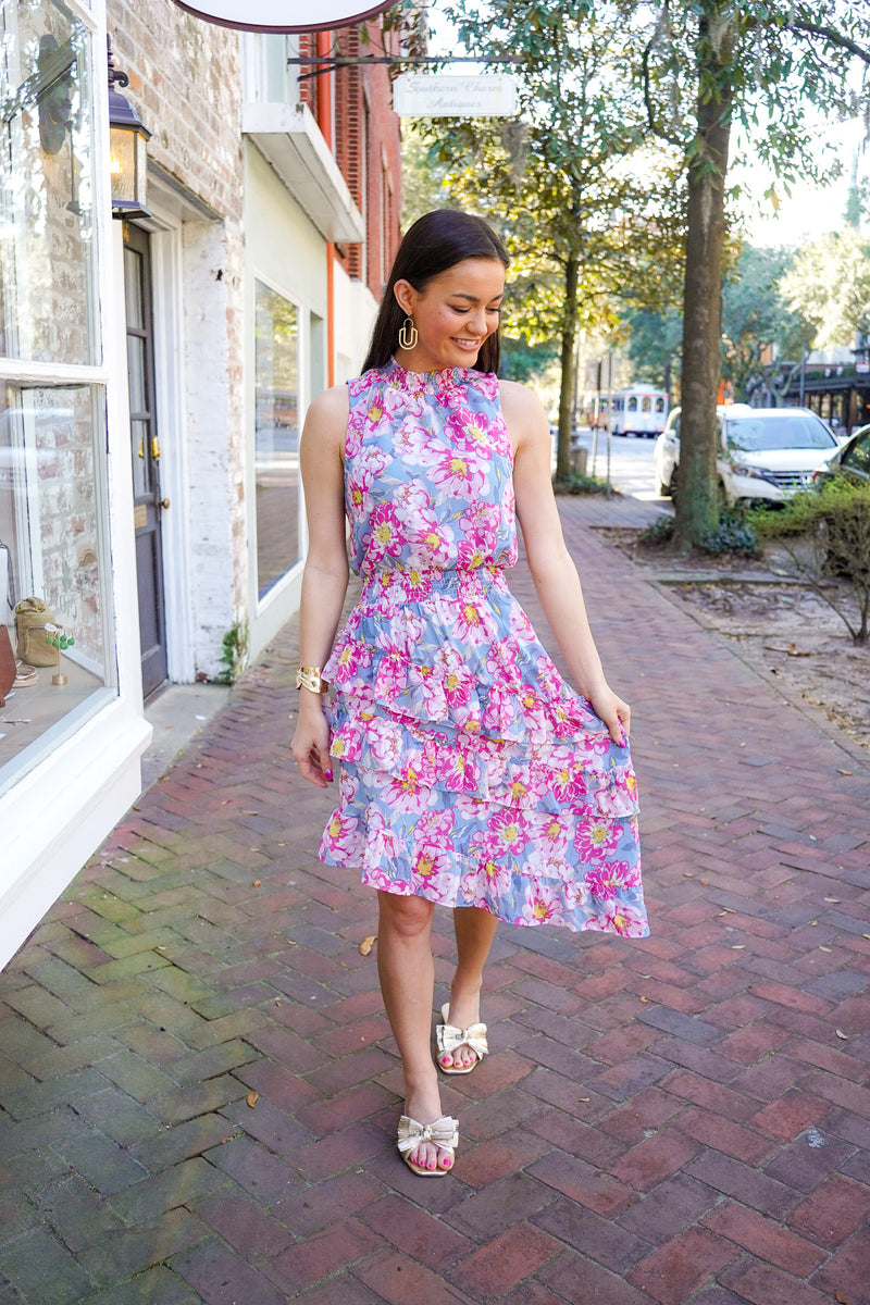 Model is wearing a dress in a floral print in blue, grey, yellow, and pink colors. It is a midi length dress has an asymmetrical skirt hem with tiered and ruffle details, and a high neckline