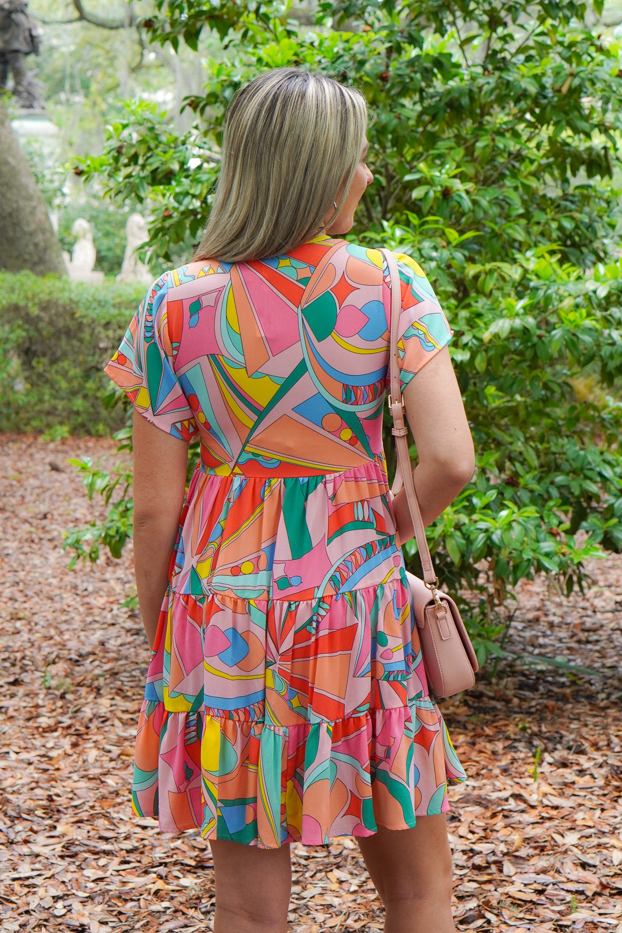 Model is wearing a multi colored mini dress with white sandals and a pink purse in front of greenery