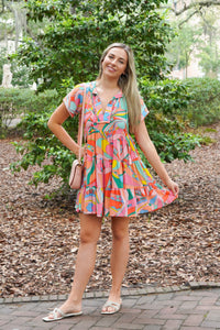 Model is wearing a multi colored mini dress with white sandals and a pink purse in front of greenery