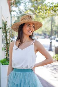 model is wearing a white satin tank paired with a blue tulle skirt and a tan sun hat.